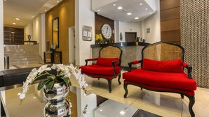 A reception area with a reception desk, two red sofas, a clock, and a vase with white flowers. - Roochelle Hotel by Nobile (Curitiba)