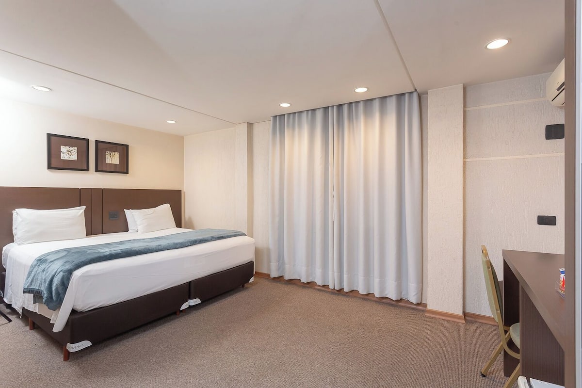 A hotel room with a bed, a desk, and a large window with curtains.