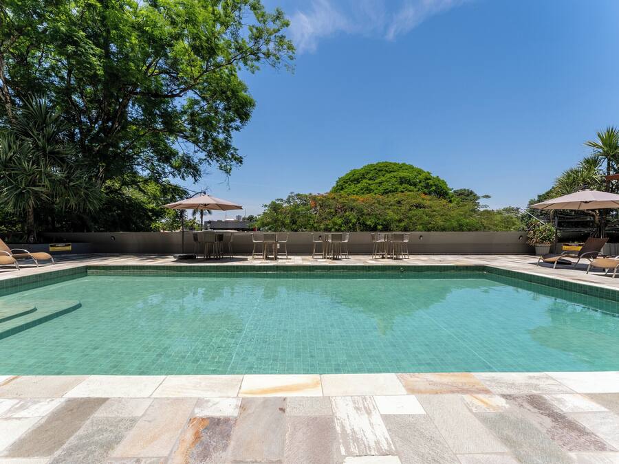 A rectangular swimming pool with tiled edges, surrounded by lounge chairs and umbrellas, with trees and a clear sky in the background.