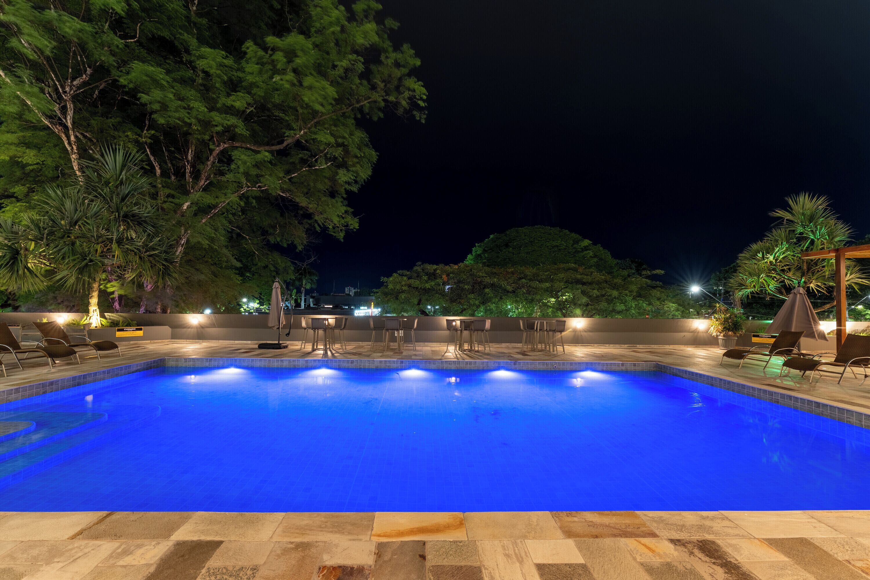 Una piscina nocturna con suelo de baldosas, rodeada de sillones de descanso y sombrillas.