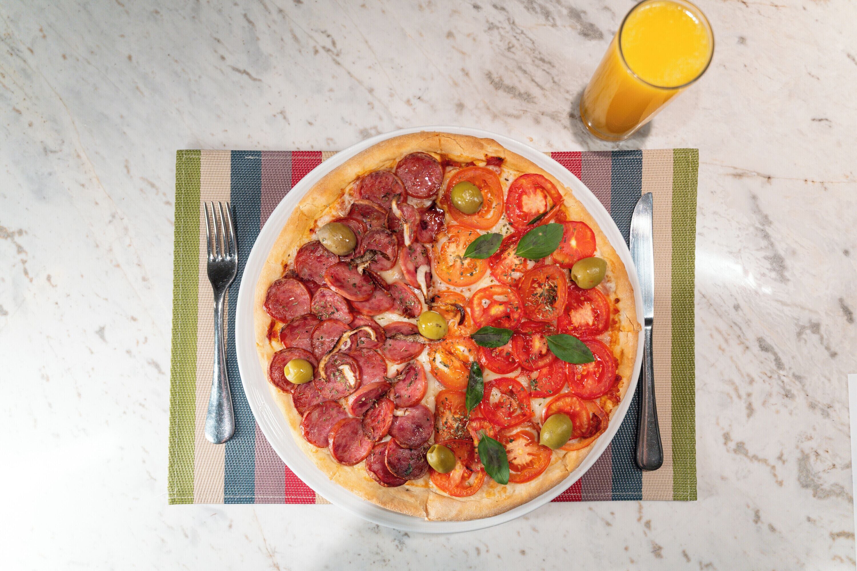 A pizza with toppings on a plate, a fork, a knife, and a glass of orange juice on a marble surface.
