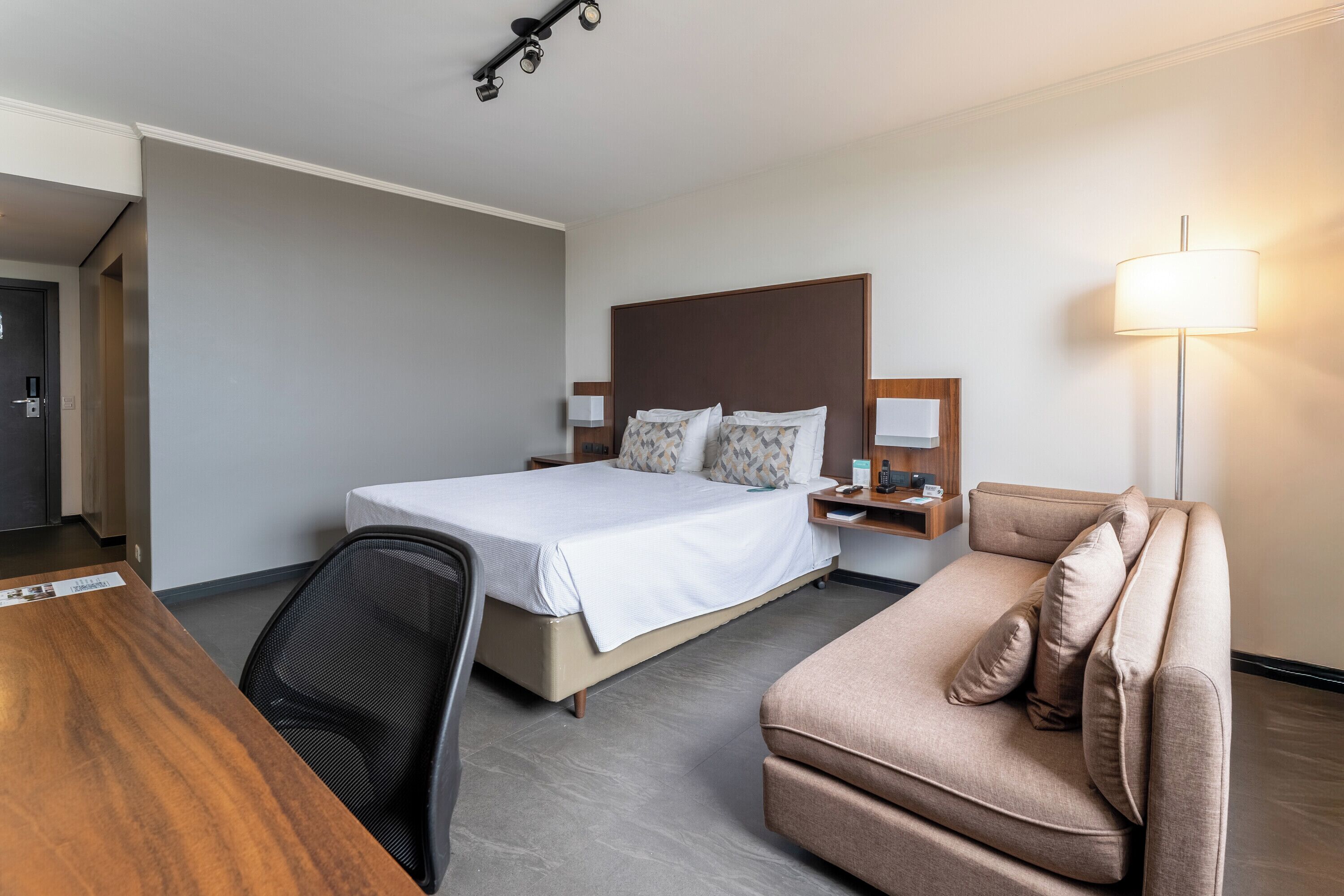 A hotel room with a bed, a desk, a chair, a sofa, a nightstand, and a lamp.