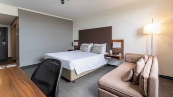 A hotel room with a bed, a desk, a chair, a sofa, a nightstand, and a lamp.