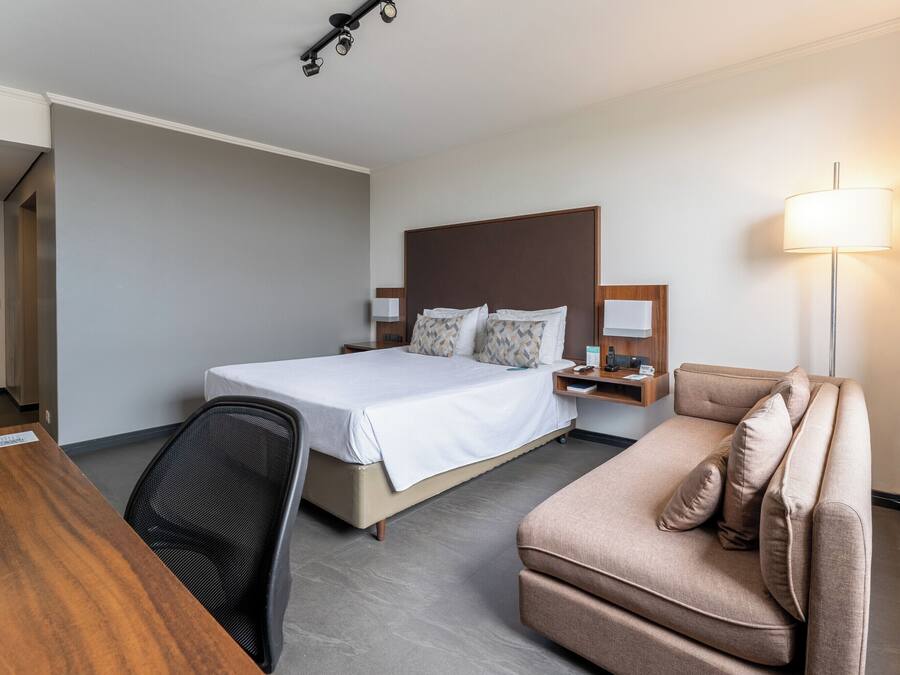 A hotel room with a bed, a desk, a chair, a sofa, a nightstand, and a lamp.