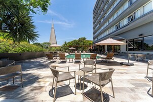 Un área de piscina al aire libre con sillones de descanso y sombrillas, rodeada de follaje y un edificio moderno.