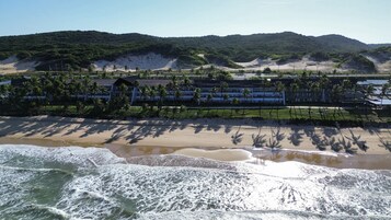 Un hotel frente al mar con vista a la playa, palmeras y montañas al fondo.