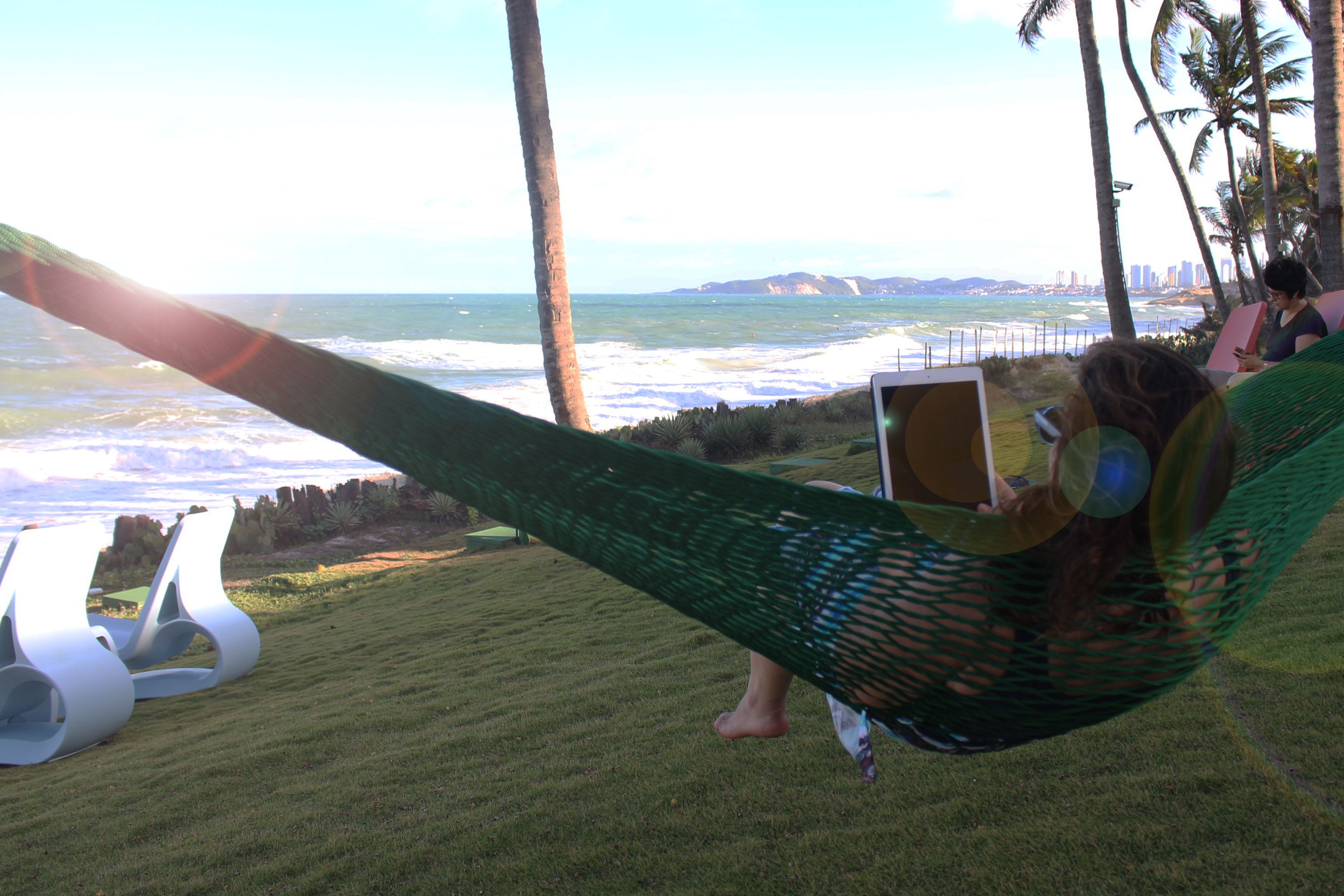 Un hamac dans lequel est assis une personne qui lit un livre, avec une vue sur une plage bordée de palmiers et, au loin, sur les contours d’une ville.