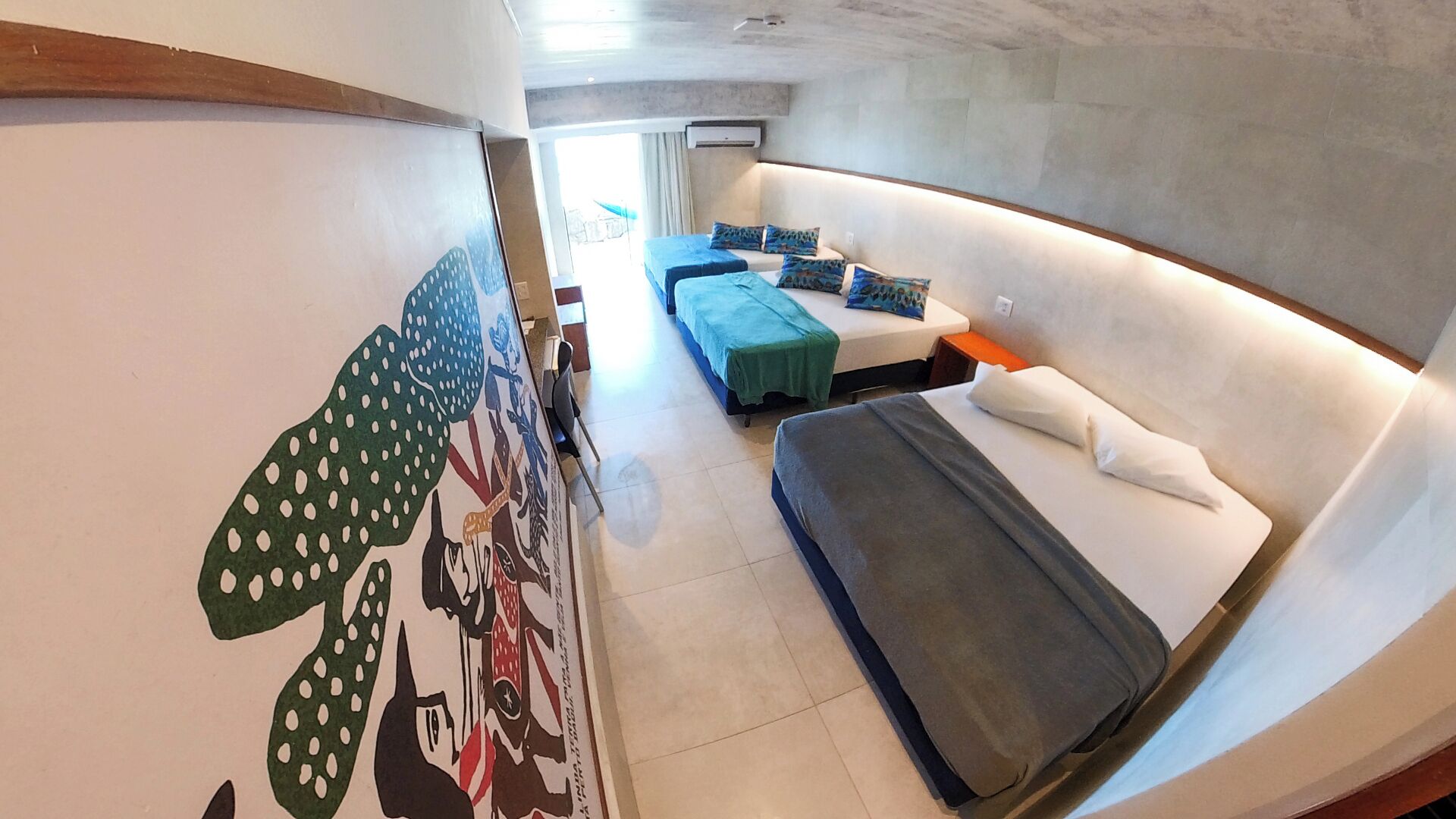 A modern hotel room with a large bed, a sofa bed, and a small table. There is a wall mural and a window with curtains.