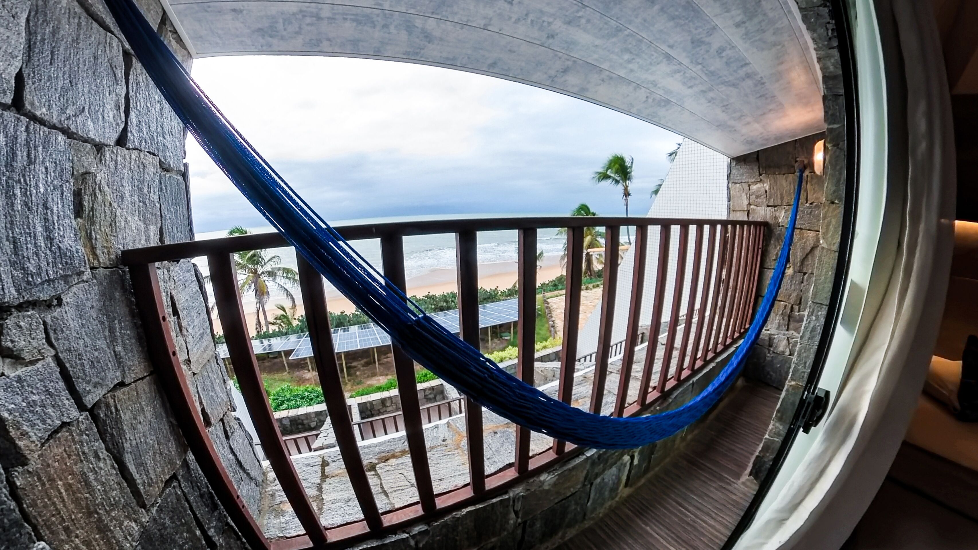 A hammock on a balcony with a view of the beach and palm trees.