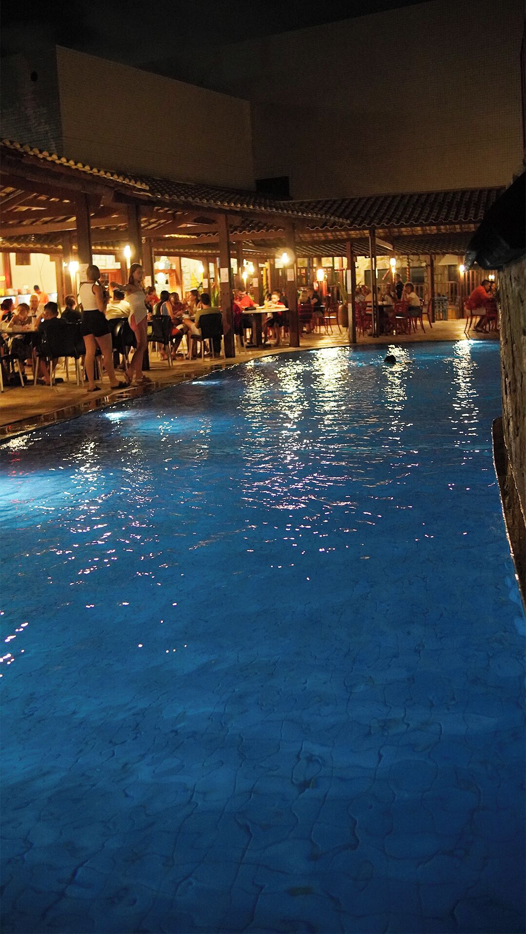 Uma piscina ao ar livre com pessoas almoçando em mesas sob um espaço coberto.