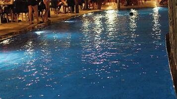 Uma piscina ao ar livre com pessoas almoçando em mesas sob um espaço coberto.