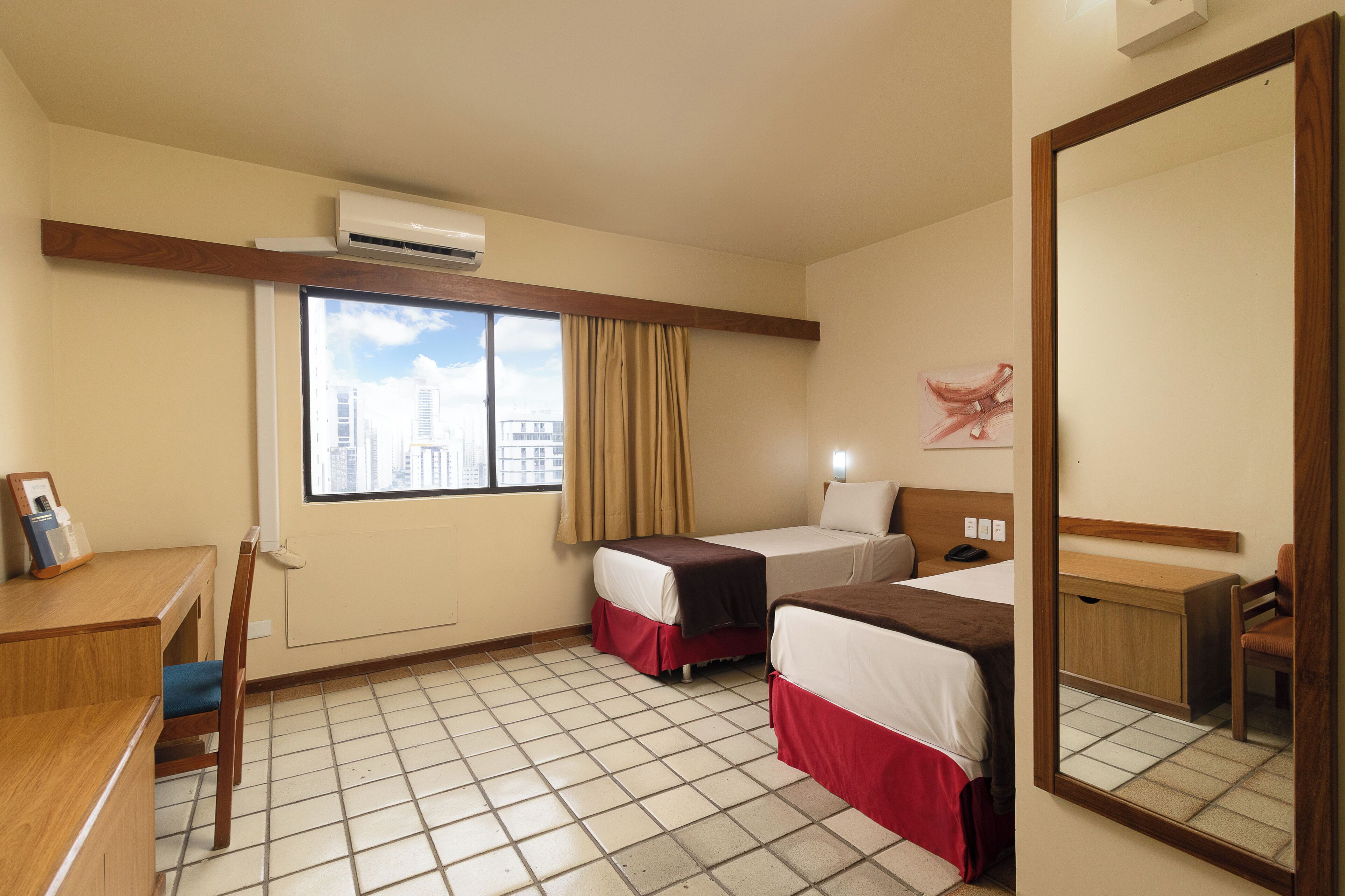 Habitación de hotel con dos camas, un escritorio, una silla, un espejo grande y una ventana con vistas a edificios.