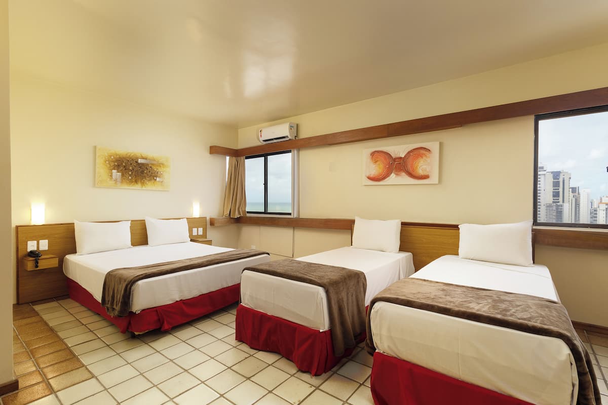 A hotel room with two beds, a window with a city view, an air conditioning unit, and artwork on the walls.