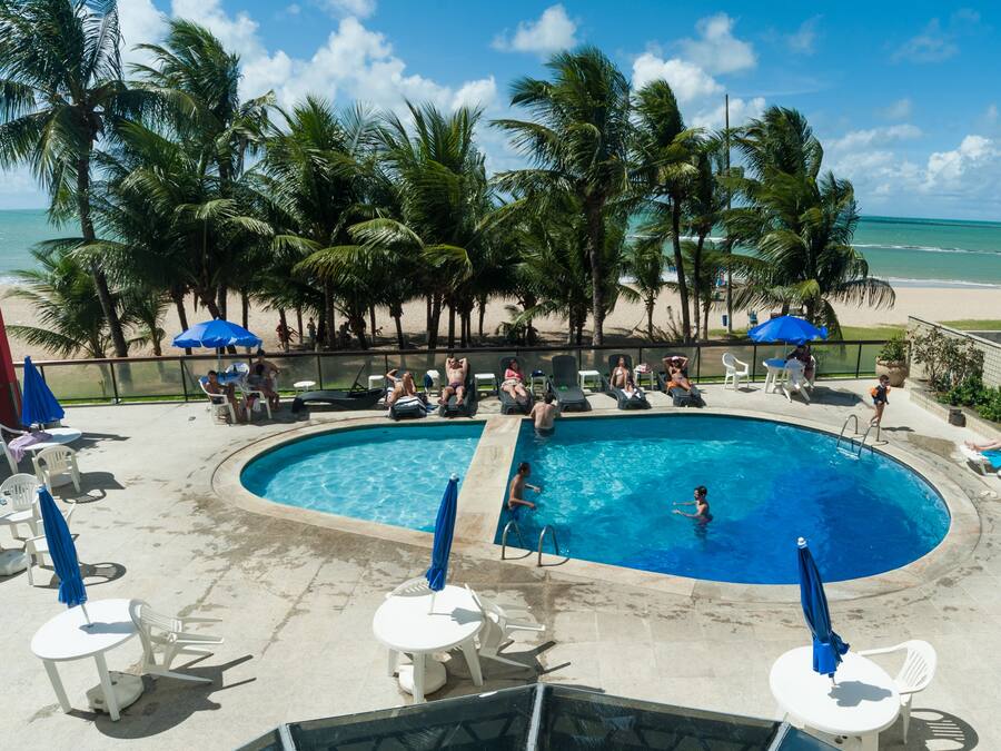 Zona de piscina con sillones de descanso y sombrillas, rodeada de palmeras y vista a la playa.
