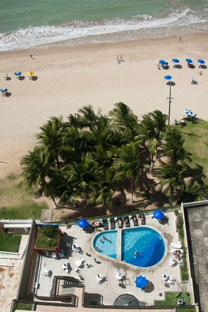 An aerial view of a beach with a pool area, palm trees, and beach umbrellas.