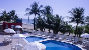 A pool area with white lounge chairs and umbrellas, surrounded by palm trees and overlooking the ocean.