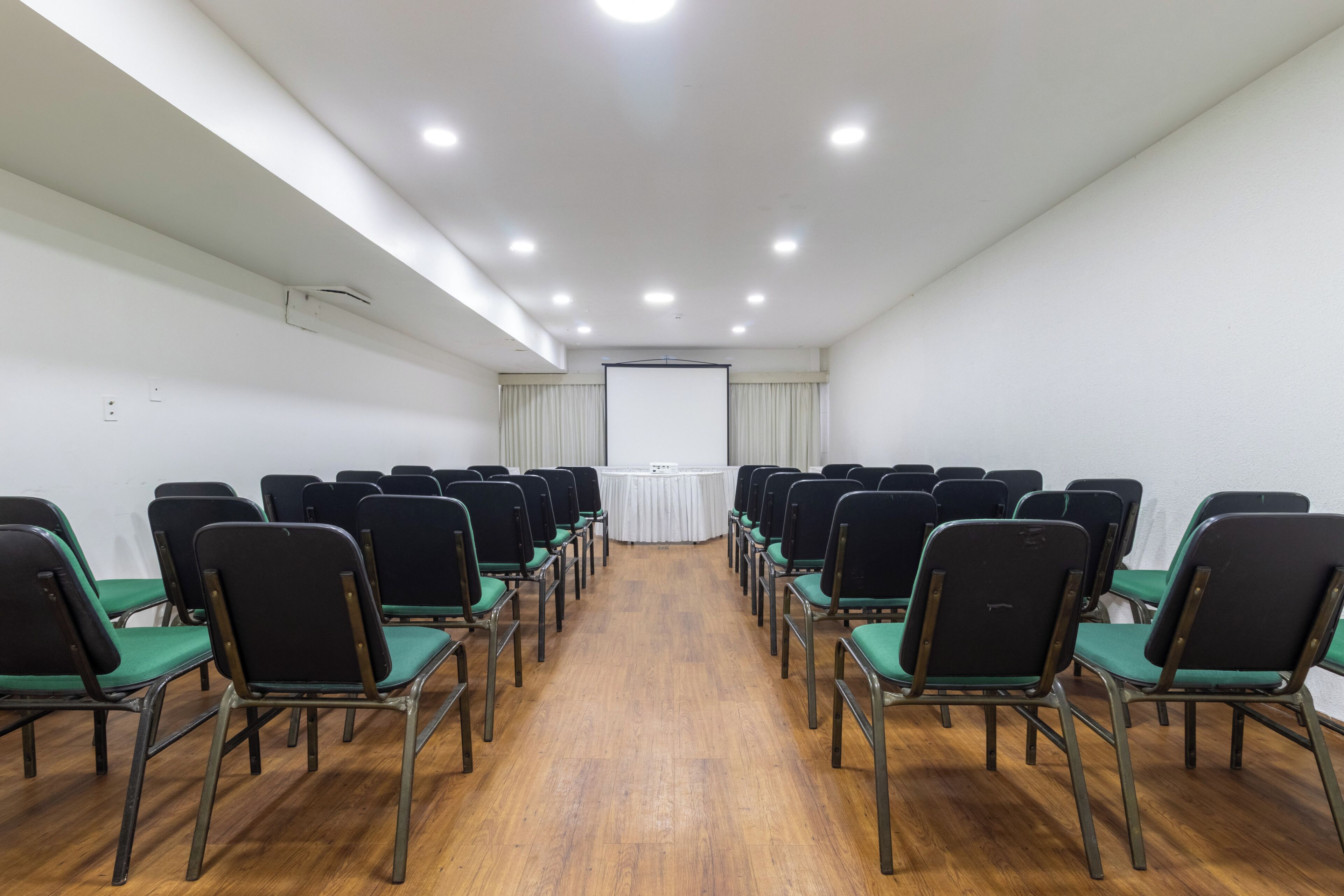 A conference room with rows of chairs, a projector screen, and a white table at the front.