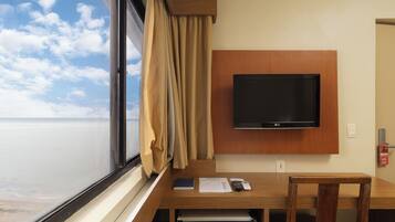 Habitación de hotel con un ventanal grande, un televisor de pantalla plana montado en la pared, un escritorio de madera con una silla y una mesita con un control remoto y un libro.