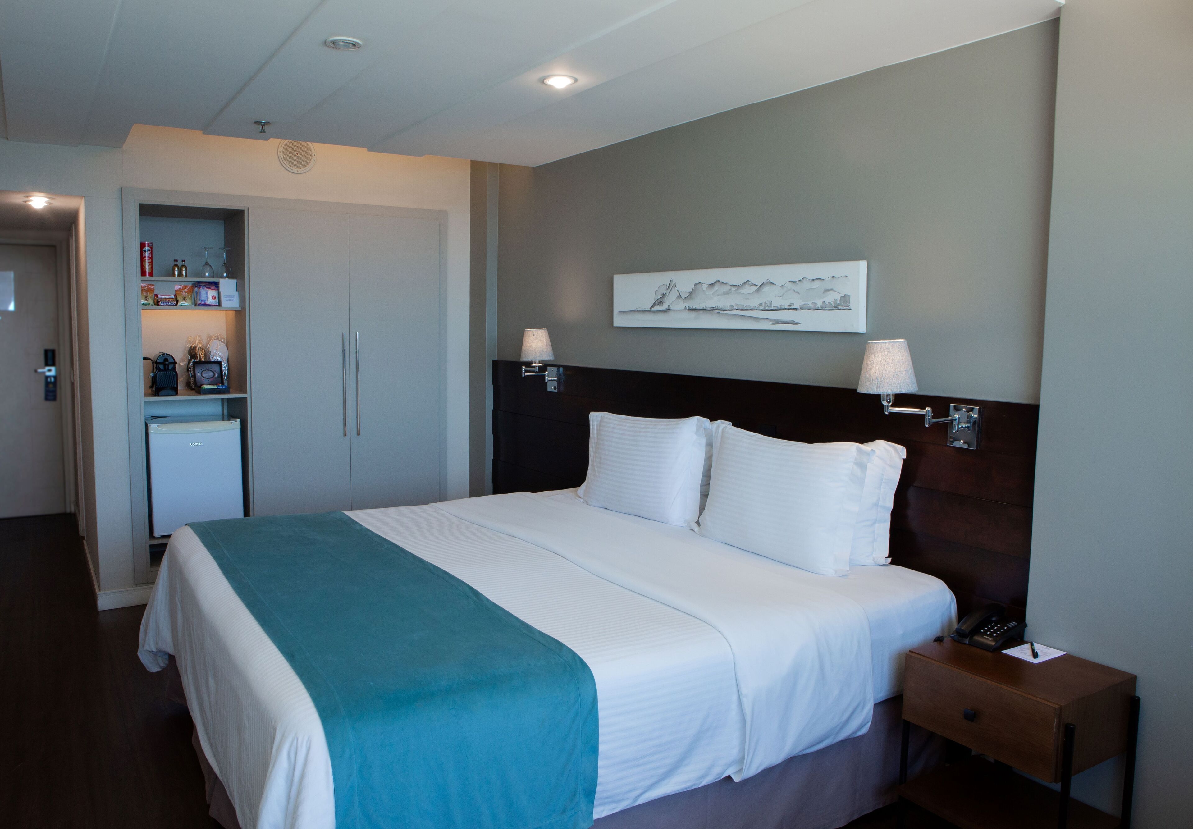 Een hotelkamer met een bed, bedlampen, een nachtkastje, een wanddecoratie en een minibar.
