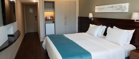 A hotel room with a large bed, a nightstand, a wall-mounted TV, a door leading to a kitchenette, and a painting on the wall.
