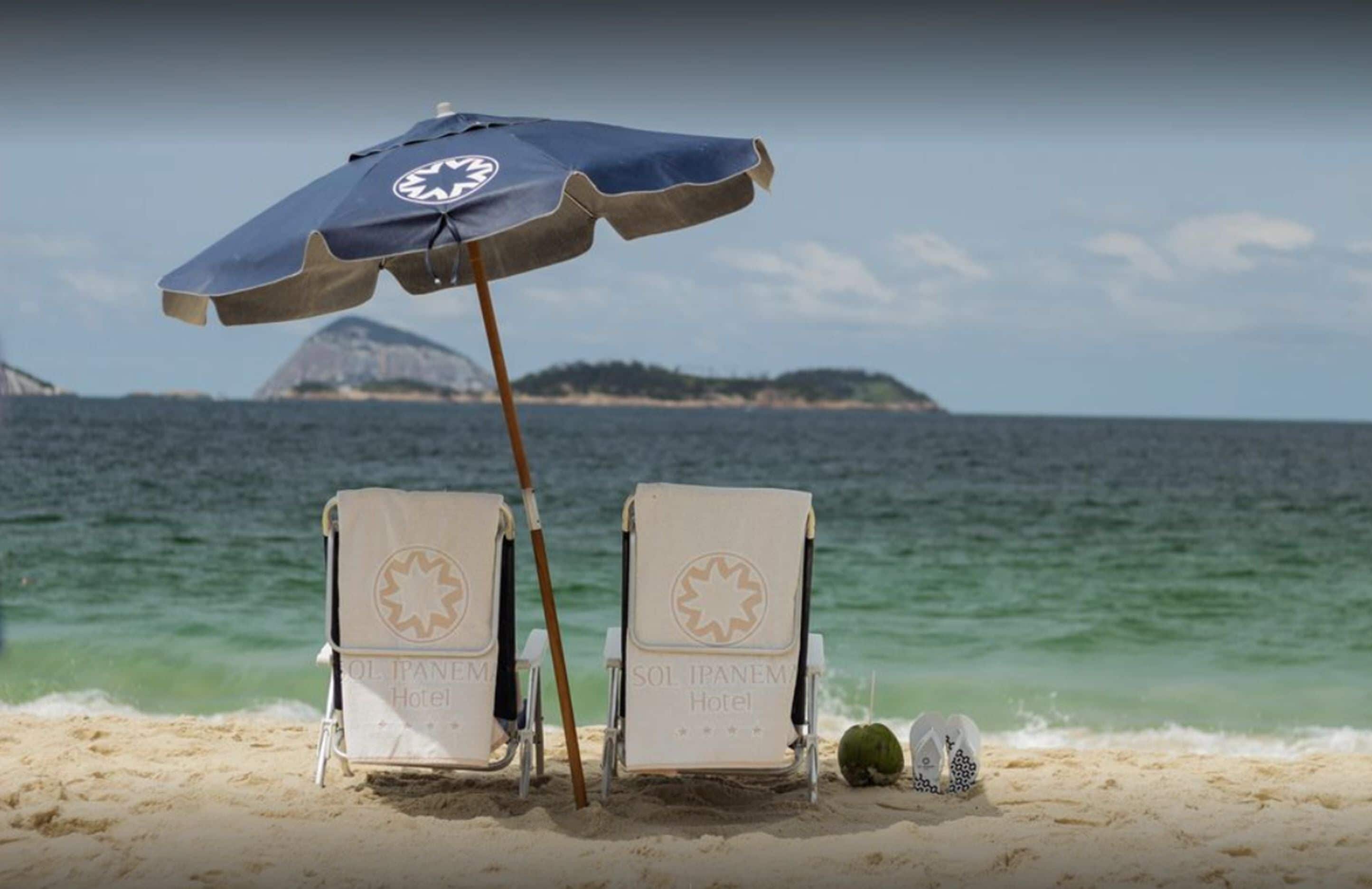 Deux chaises de plage, une serviette, un parasol et une noix de coco, le tout sur une plage de sable avec vue sur l’océan et une île lointaine.