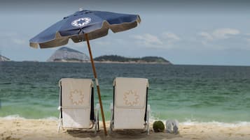 Deux chaises de plage, une serviette, un parasol et une noix de coco, le tout sur une plage de sable avec vue sur l’océan et une île lointaine.