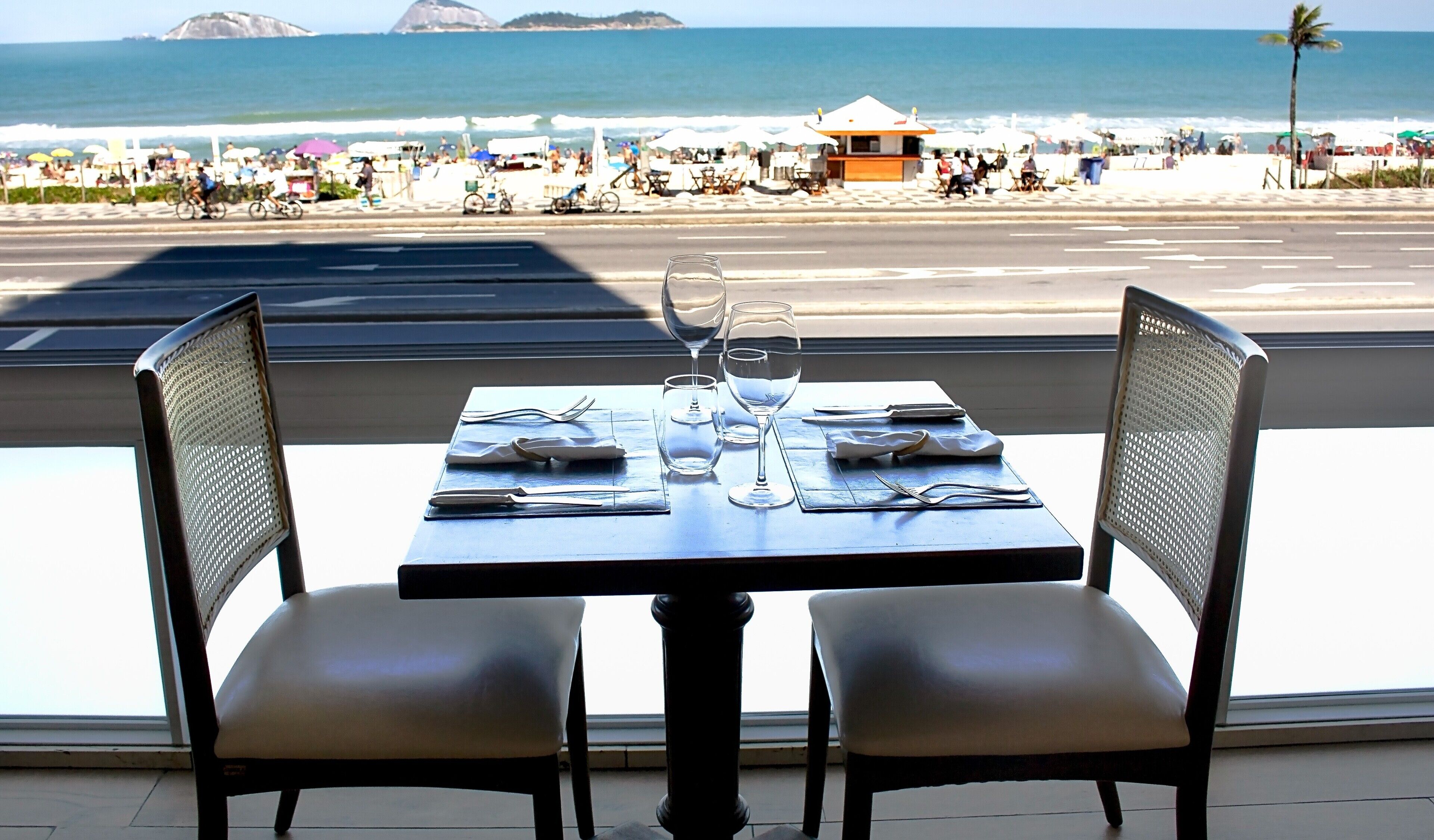 A dining table with chairs, set for two, overlooking a beach with umbrellas and people.