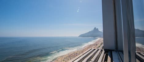 Vista de la playa desde un punto elevado, mostrando una extensa franja de arena con sombrillas y gente, una montaña al fondo y un cielo despejado.