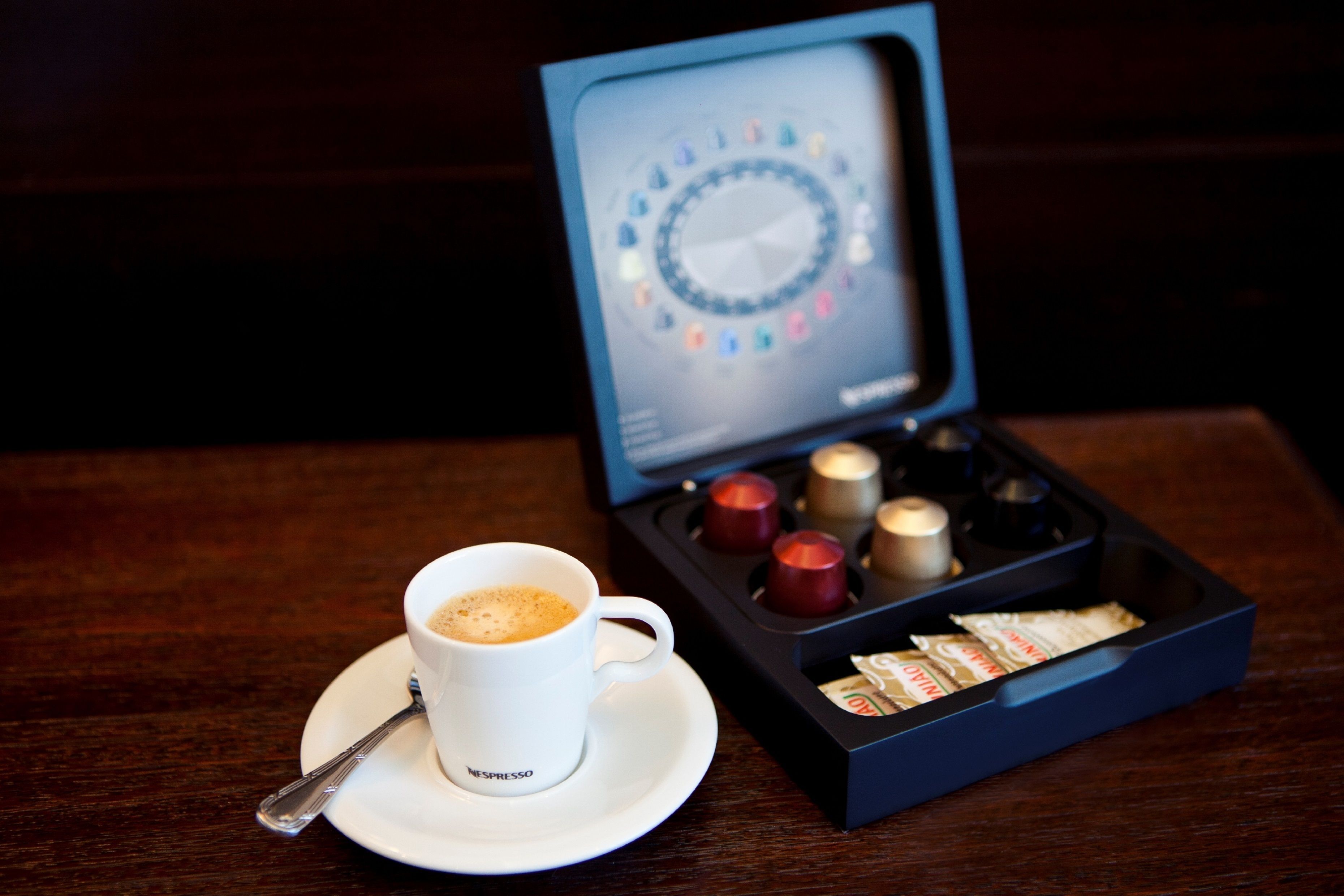 Une tasse de café avec une cuillère sur une soucoupe, une machine à café et une boîte de capsules de café sur une table en bois.