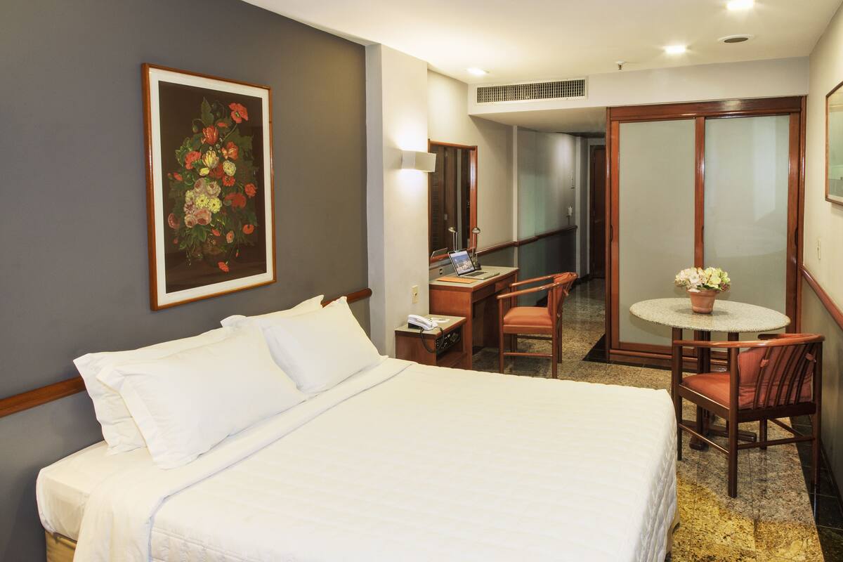 A hotel room with a bed, a desk, a chair, a small table with a flower, and a painting on the wall.