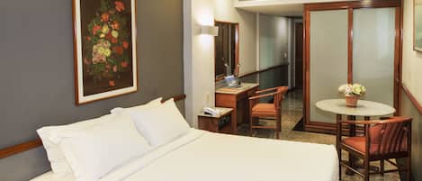 A hotel room with a bed, a desk, a chair, a small table with a flower, and a painting on the wall.