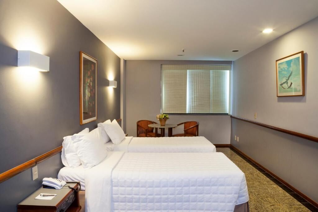 Habitación de hotel con dos camas, una pequeña mesa con sillas, una ventana con persianas y obras de arte en las paredes.
