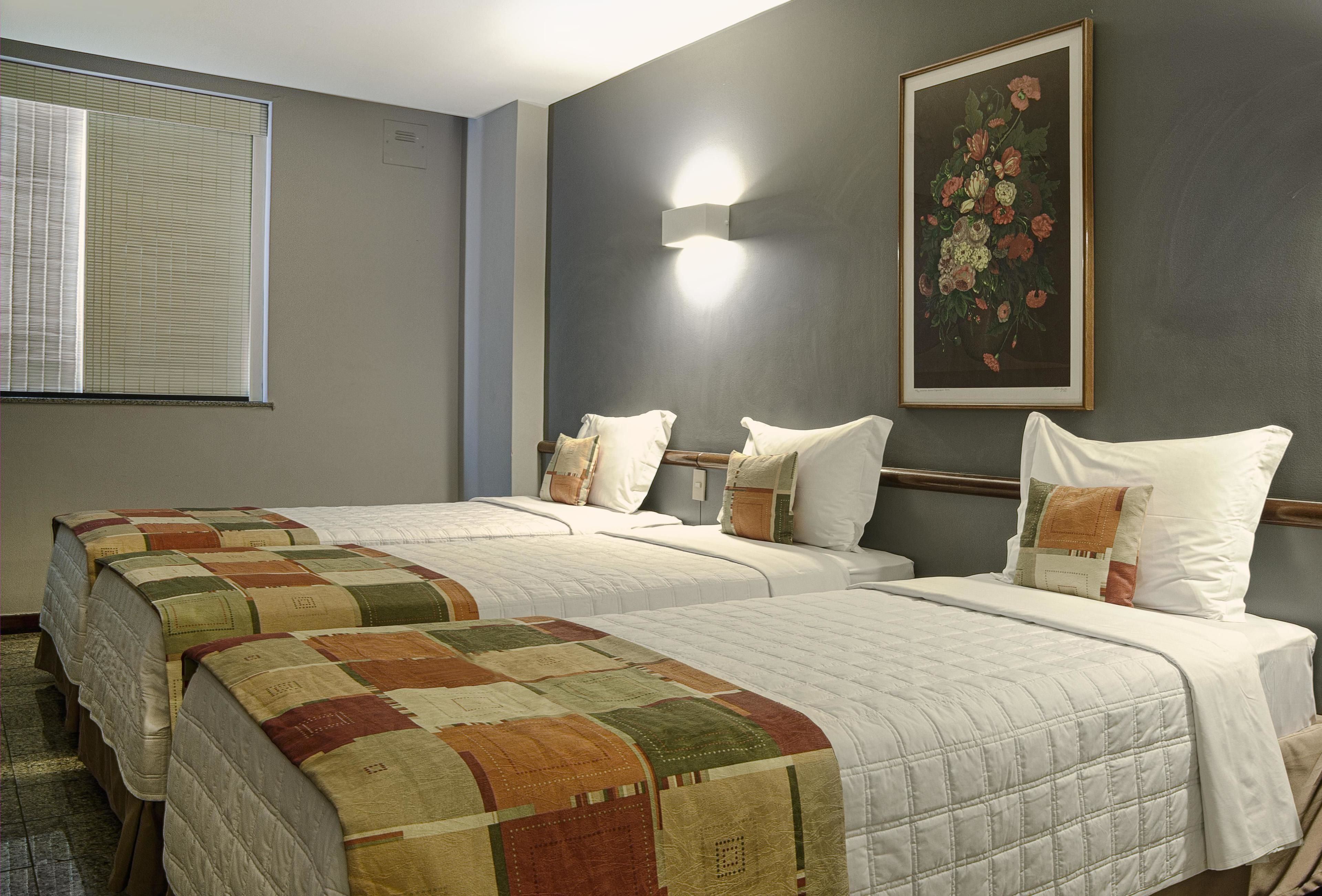 A hotel room with two beds, a large window with blinds, a wall-mounted light, and a floral painting.