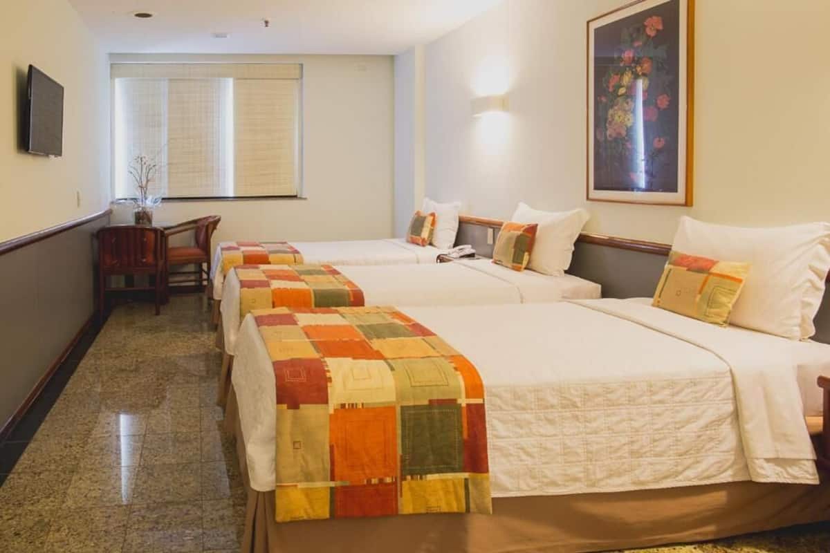 A hotel room with two beds, a desk, and a chair. There is a window with blinds, a painting on the wall, and a television mounted on the wall.
