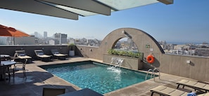 Une piscine sur le toit offrant une vue sur la ville, des chaises longues et une petite cascade décorative.
