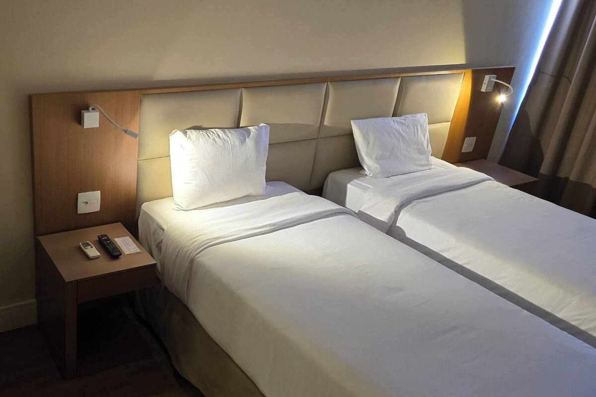 Une chambre d’hôtel avec un lit simple, une table de chevet avec un téléphone et une télécommande, et un petit bureau avec une chaise.