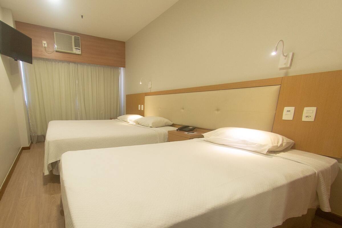 A hotel room with two beds, a television, an air conditioning unit, a bedside telephone, and a window with curtains.