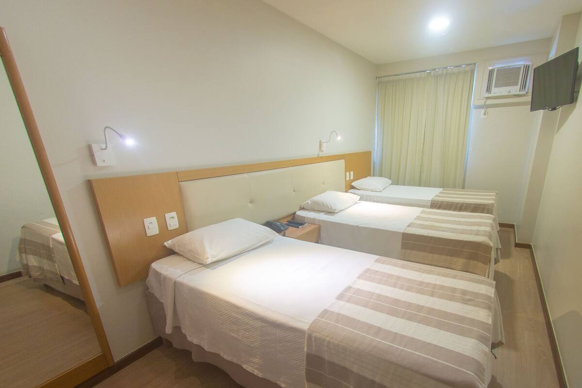 A hotel room with three single beds, a wooden headboard, a bedside table with a lamp, and a television mounted on the wall.