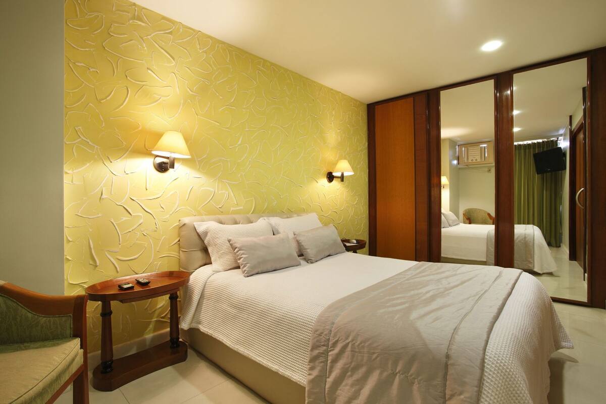 A hotel room with a bed, bedside tables, a mirror, and a green chair.