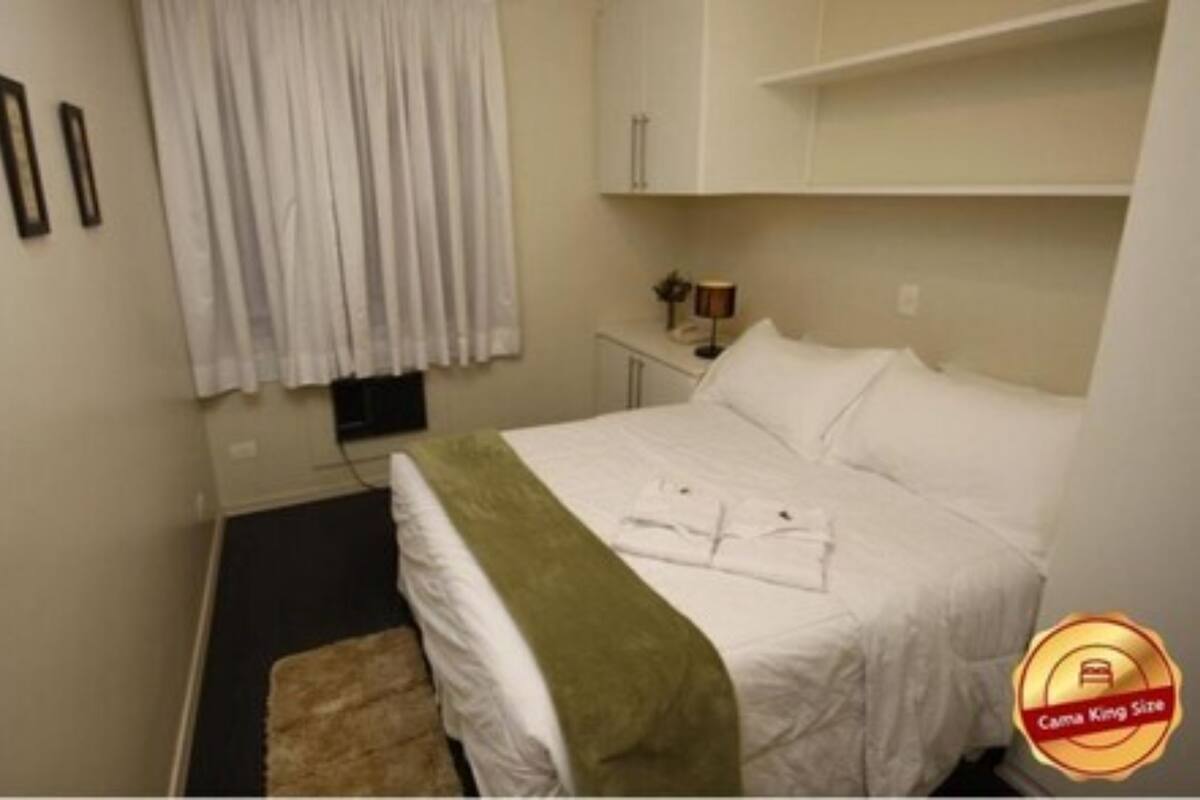 A hotel room with a bed, a small bedside table with a lamp, a window with curtains, and a wall-mounted shelf.