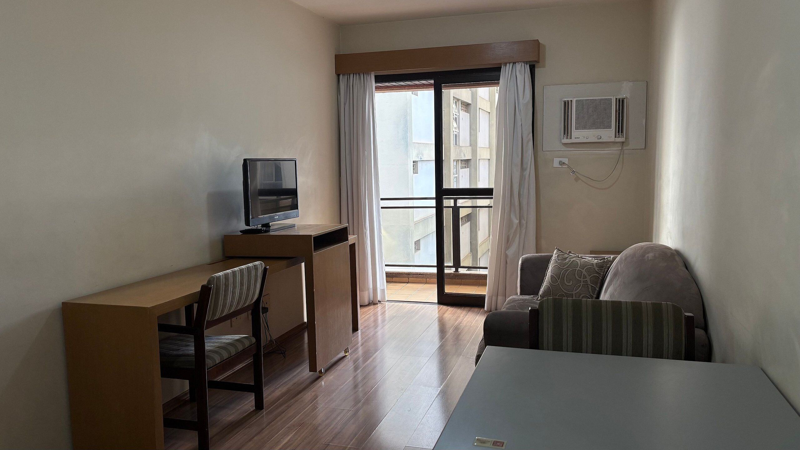A hotel room with a desk, chair, television, and a balcony with curtains.
