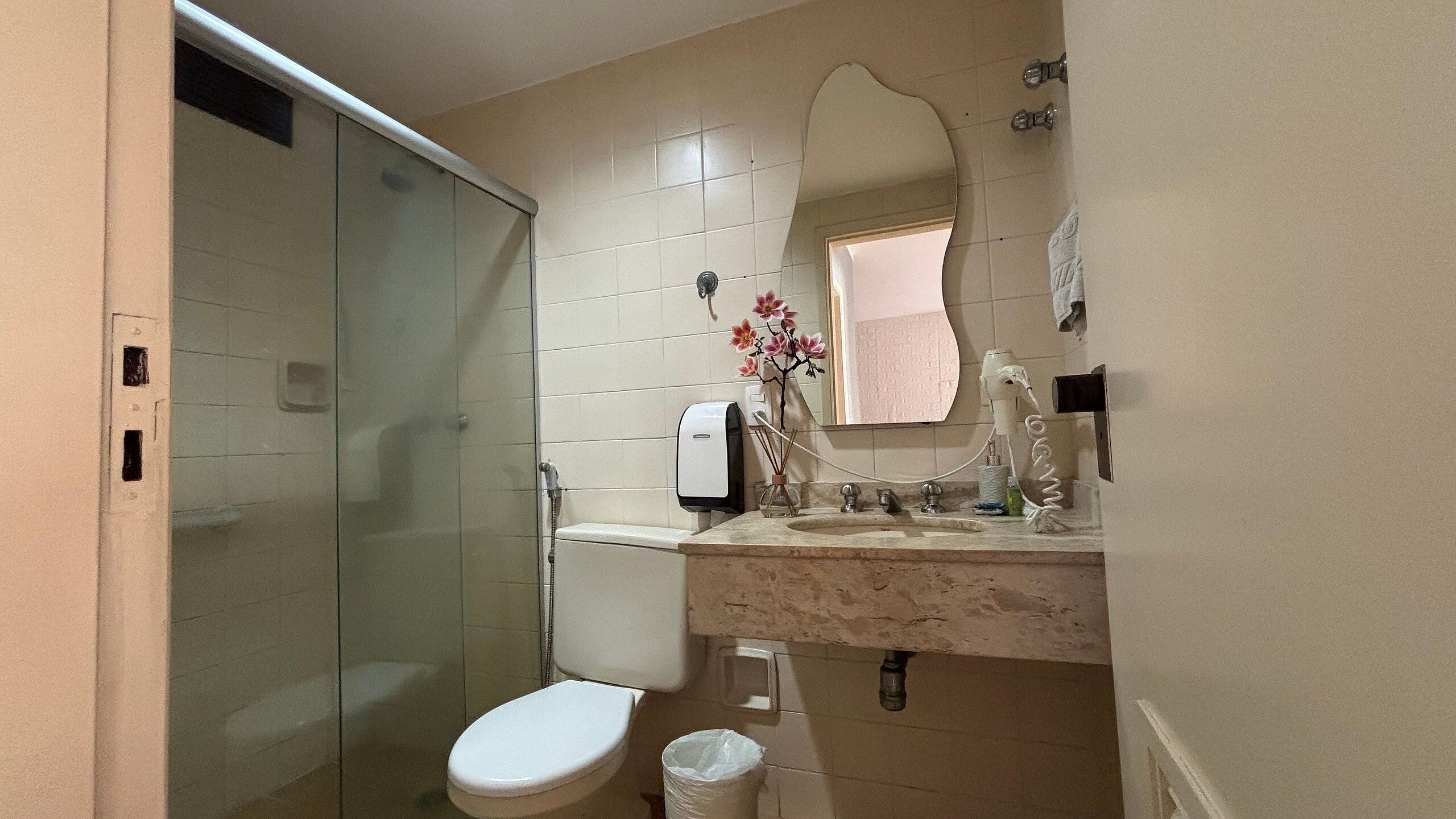 A bathroom with a glass shower enclosure, a toilet, a sink with a mirror, and a small window.