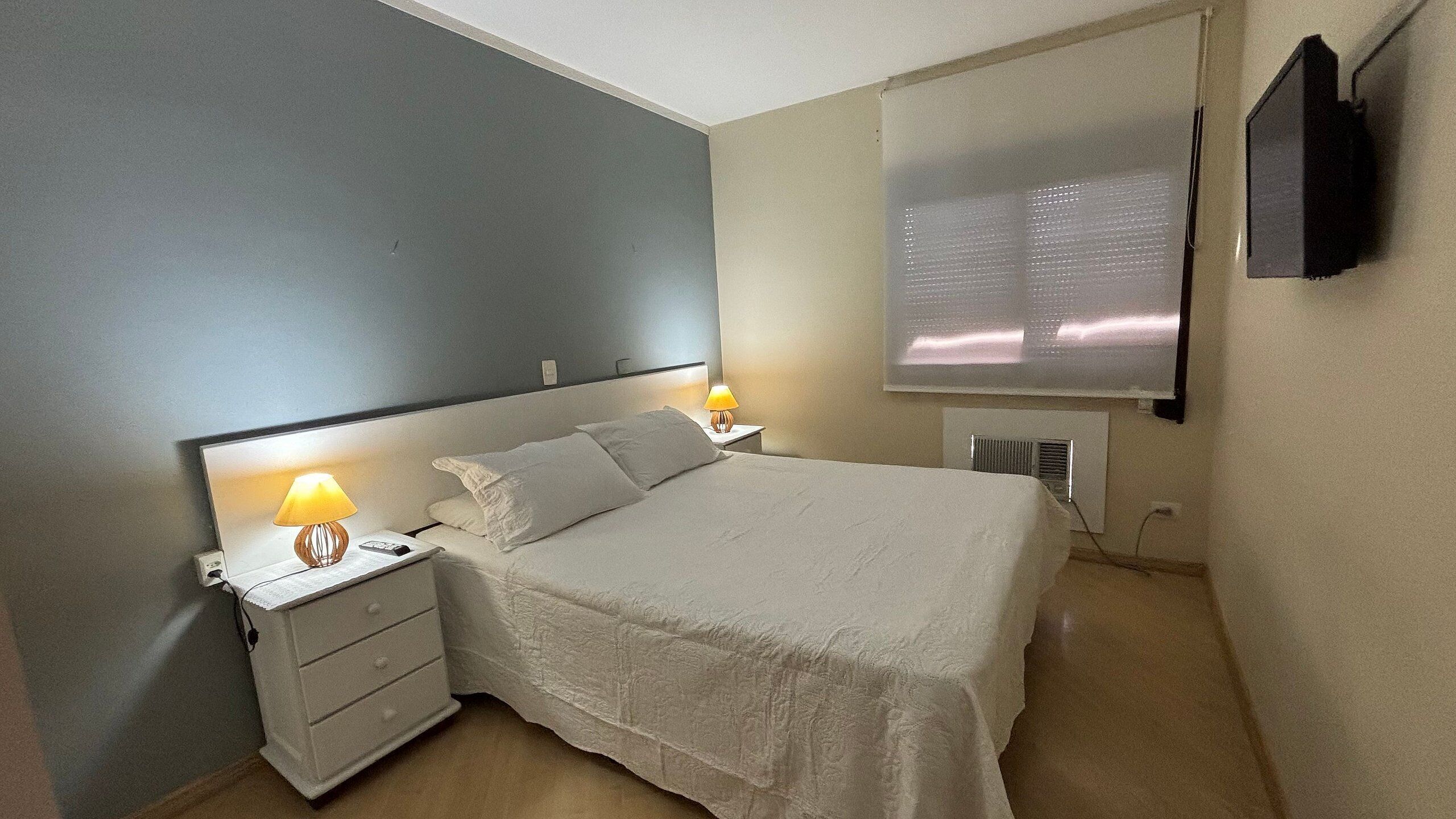 Un dormitorio con cama, mesitas de noche, lámparas, un televisor y una ventana con persianas.