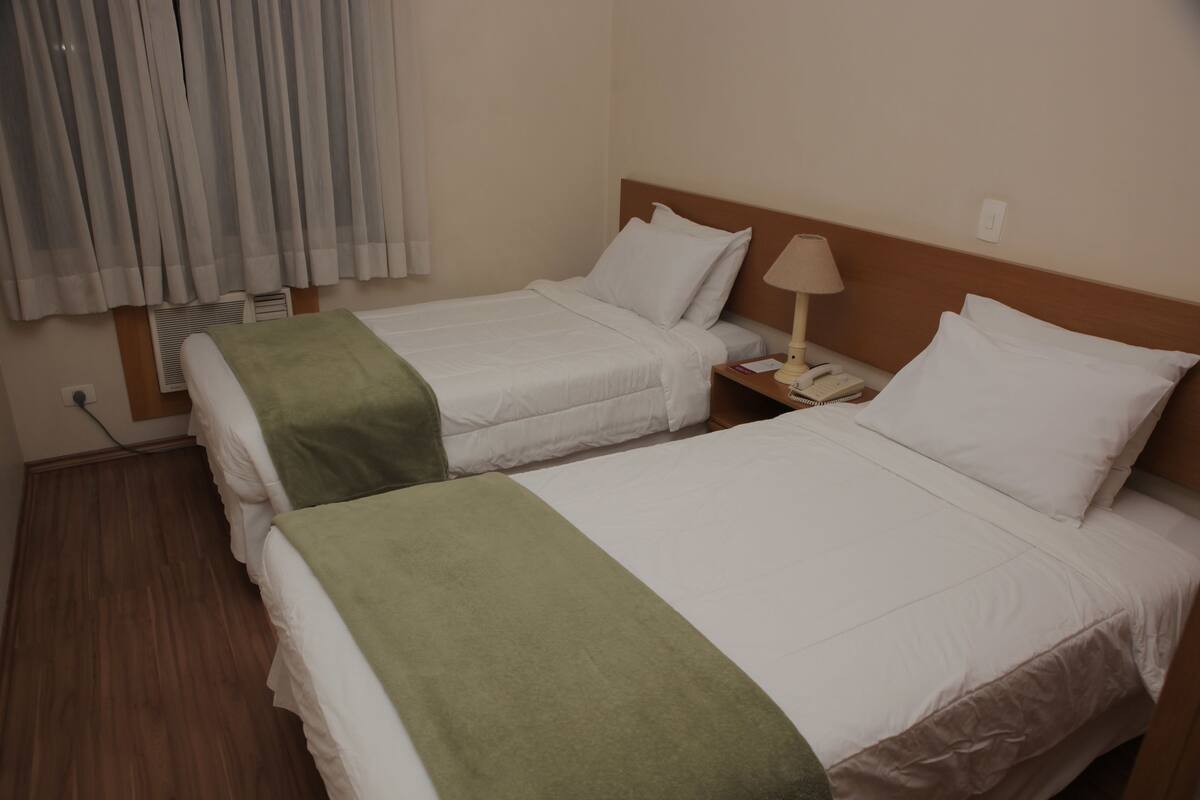 A hotel room with two single beds, a nightstand with a lamp, and a window with curtains.