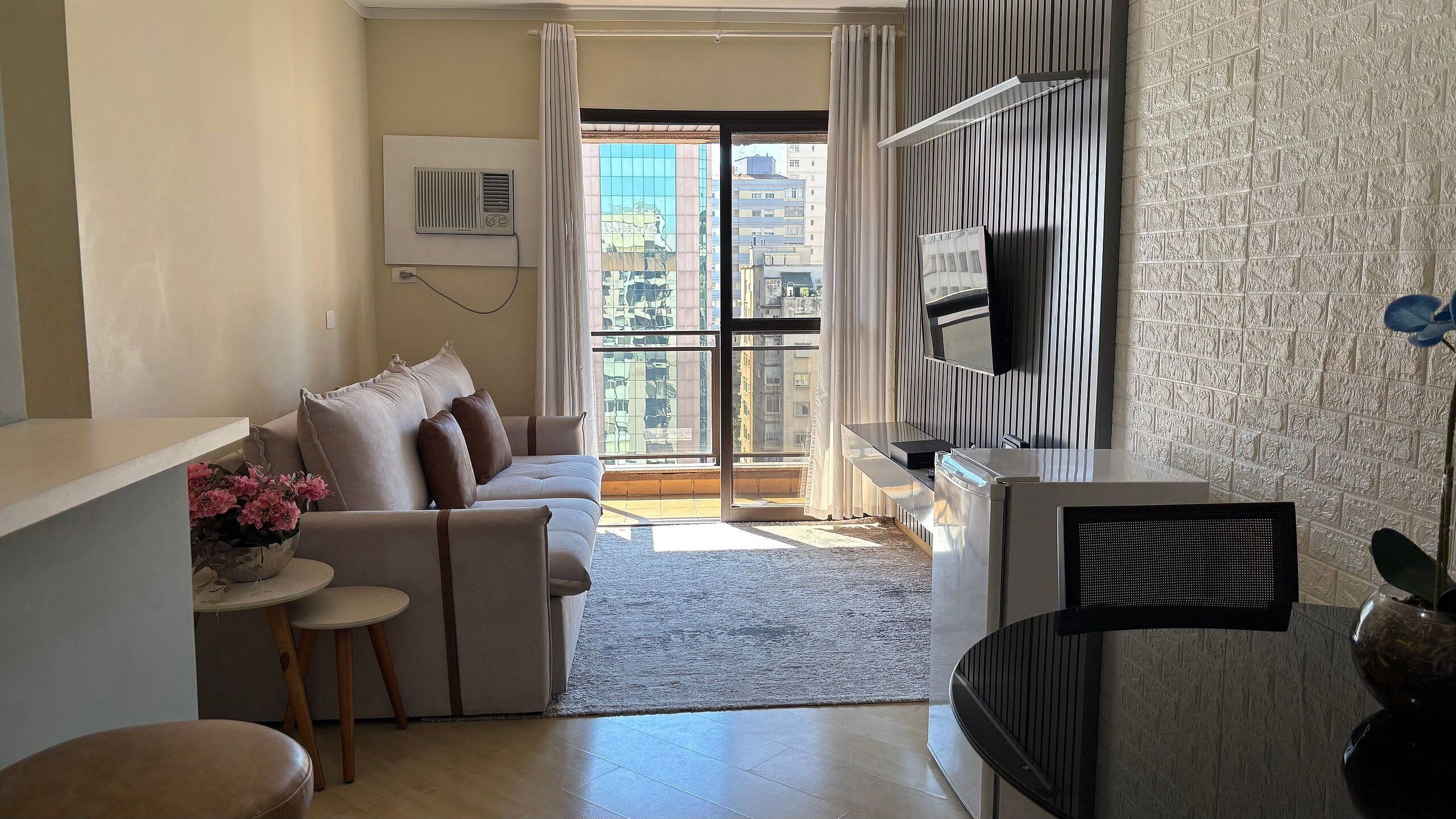 A modern living room with a sofa, a small table with flowers, a TV, and a view of the city through large windows.