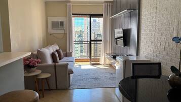 A modern living room with a sofa, a small table with flowers, a TV, and a view of the city through large windows.