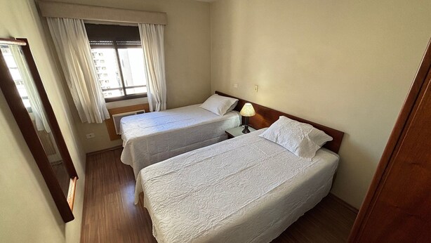 A hotel room with two single beds, a nightstand with a lamp, a window with curtains, and a wooden floor. - London Class Hotels (São Paulo)