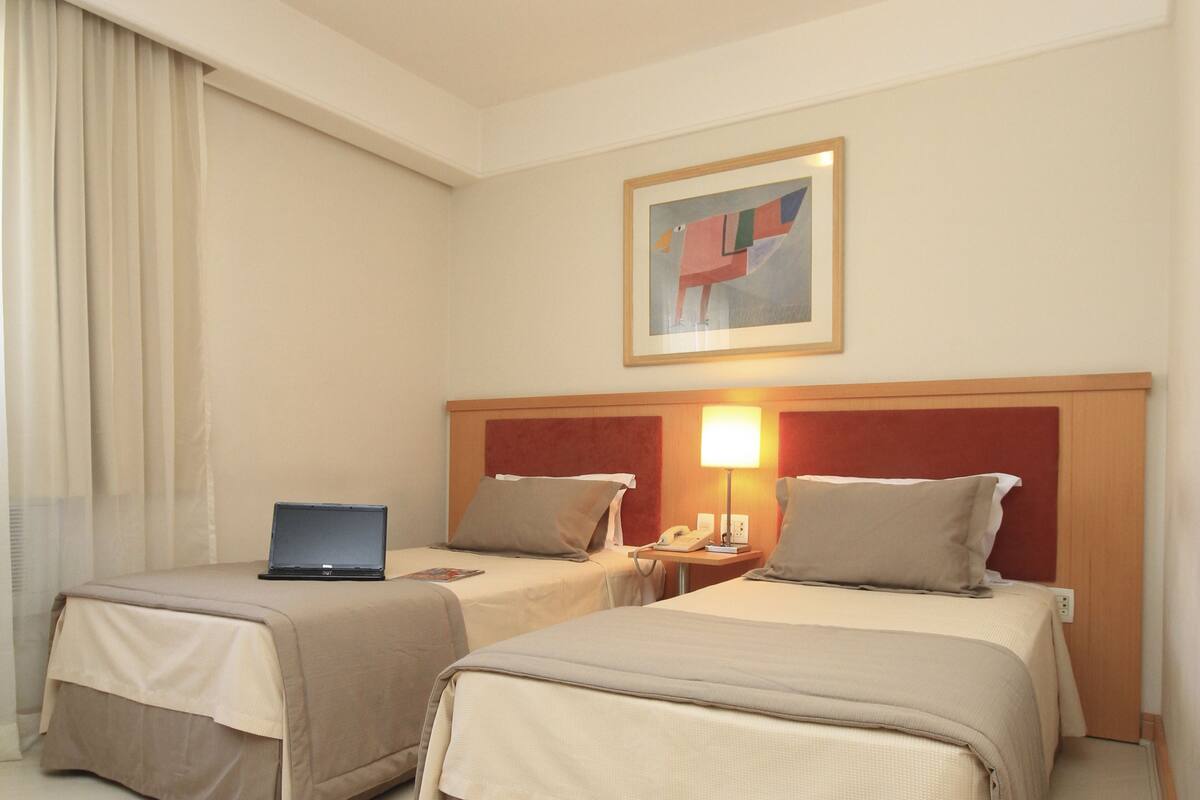 Een hotelkamer met twee bedden, een bureau met een laptop, een bedlamp en een schilderij aan de muur.