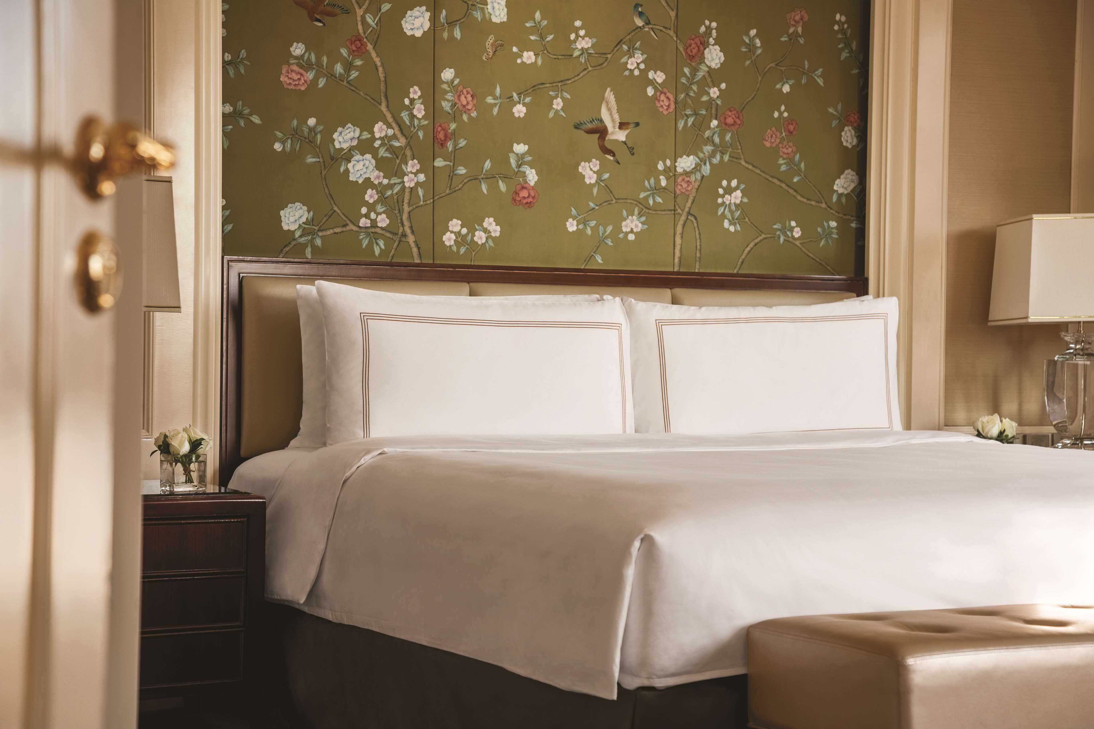 Een net opgemaakt bed met witte lakens, een hoofdbord met bloemen behang, een nachtkastje met een lamp en een kleine vaas met bloemen.