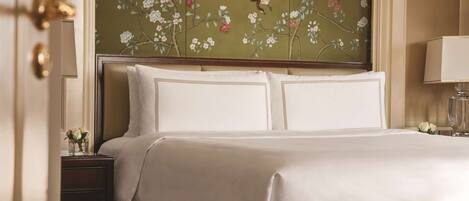 A neatly made bed with white linens, a floral wallpaper headboard, a bedside table with a lamp, and a small vase with flowers.